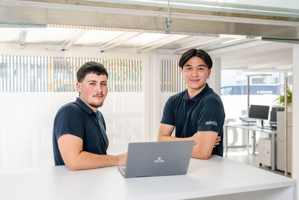 Two professionals working on a laptop at a modern white desk in an office setting.