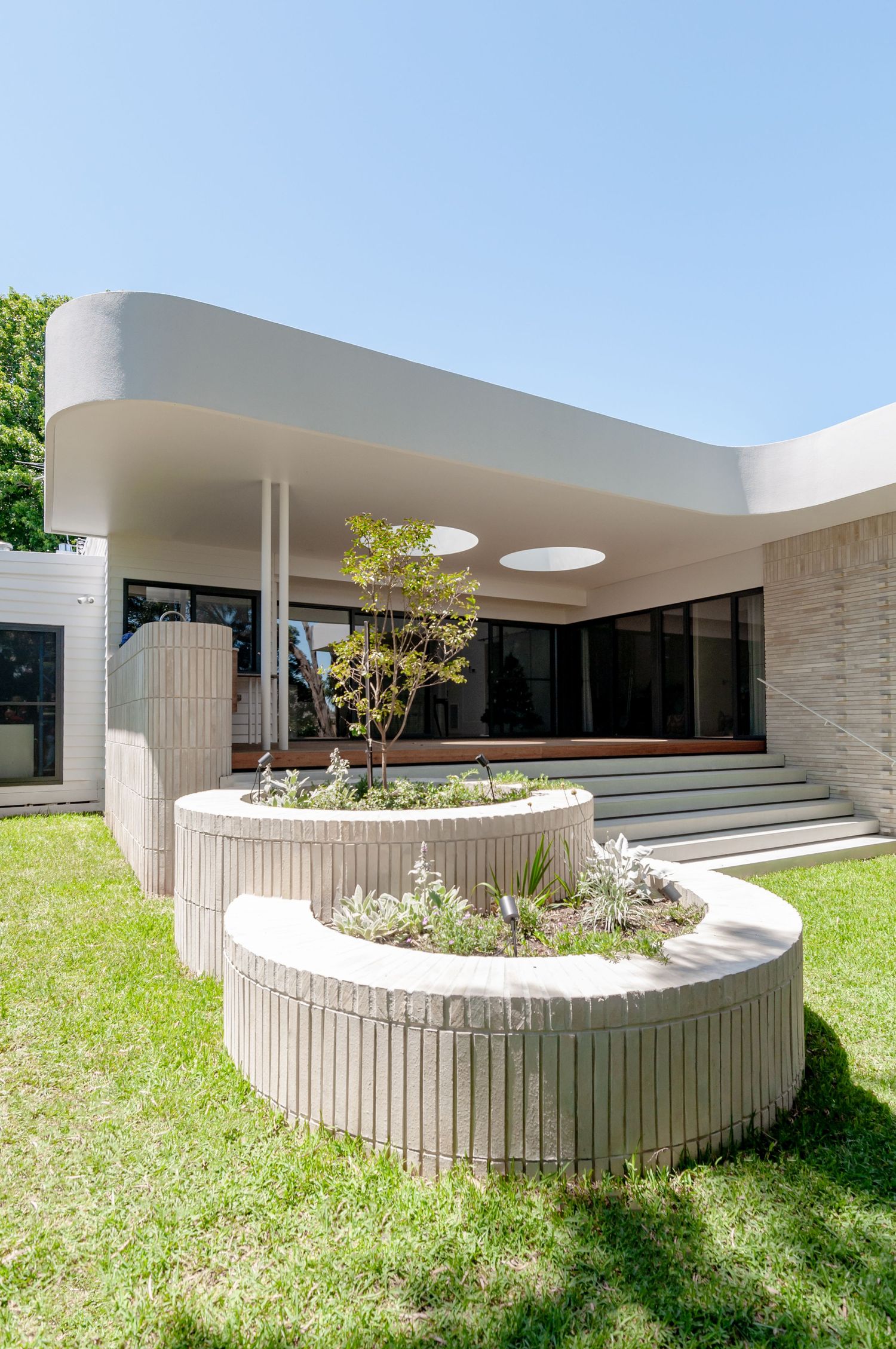 Modern home exterior with curved planters and steps leading to glass entrance under white overhang with skylight details.