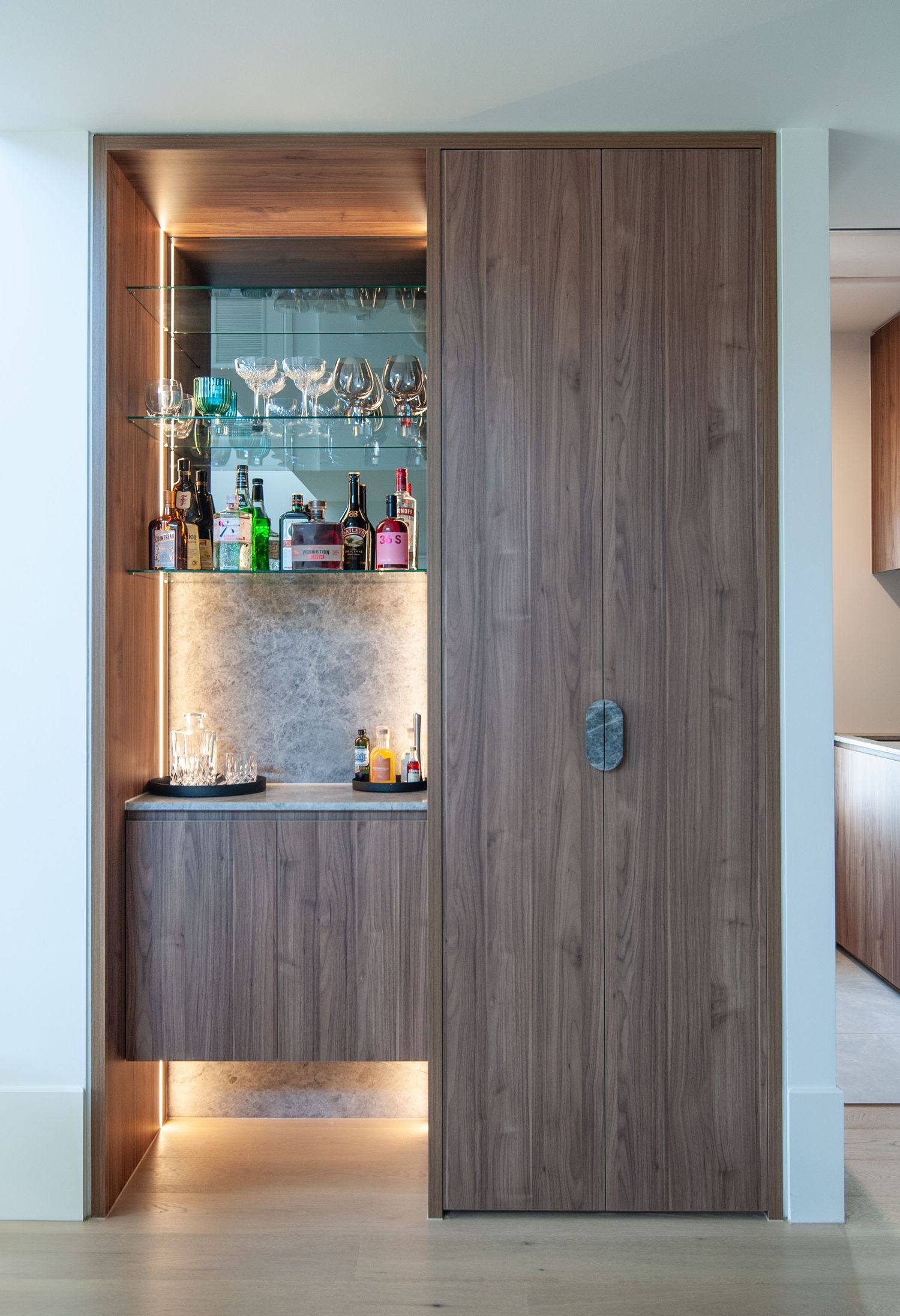 Modern built-in bar nook with wood paneling, glass shelving, and ambient lighting displays liquor bottles and glassware.