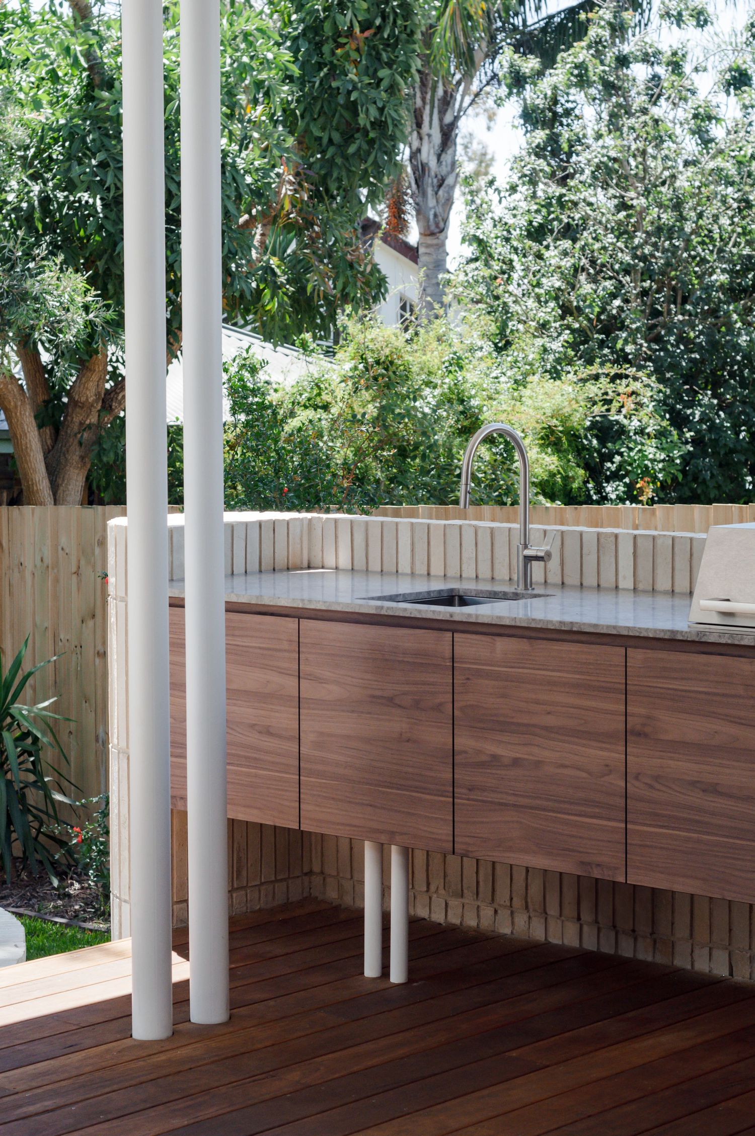 Outdoor kitchen cabinetry with sink and stainless steel faucet surrounded by lush greenery.