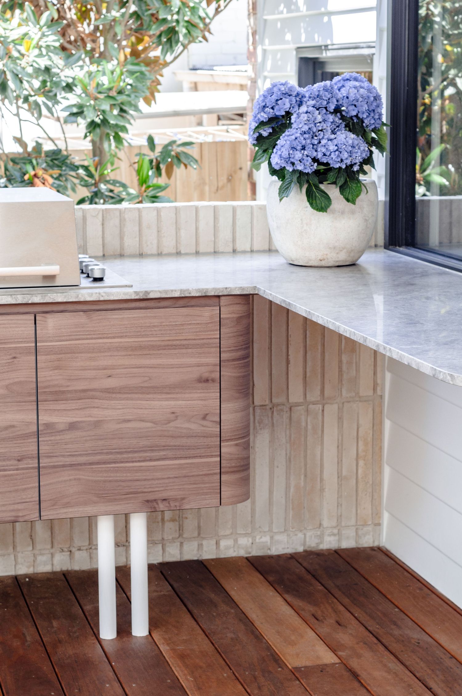 Stylish outdoor kitchen with white marble countertop and potted blue hydrangeas against wooden panels.