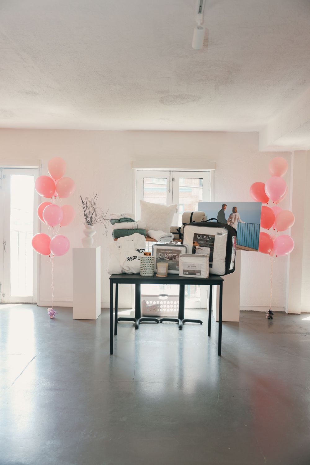 Pink balloons decorate a minimalist room with a black table display setup near windows.