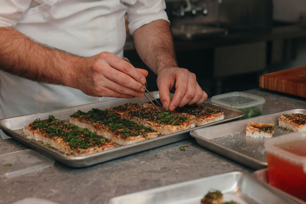 Hands preparing food on metal baking sheets in professional kitchen setting.
