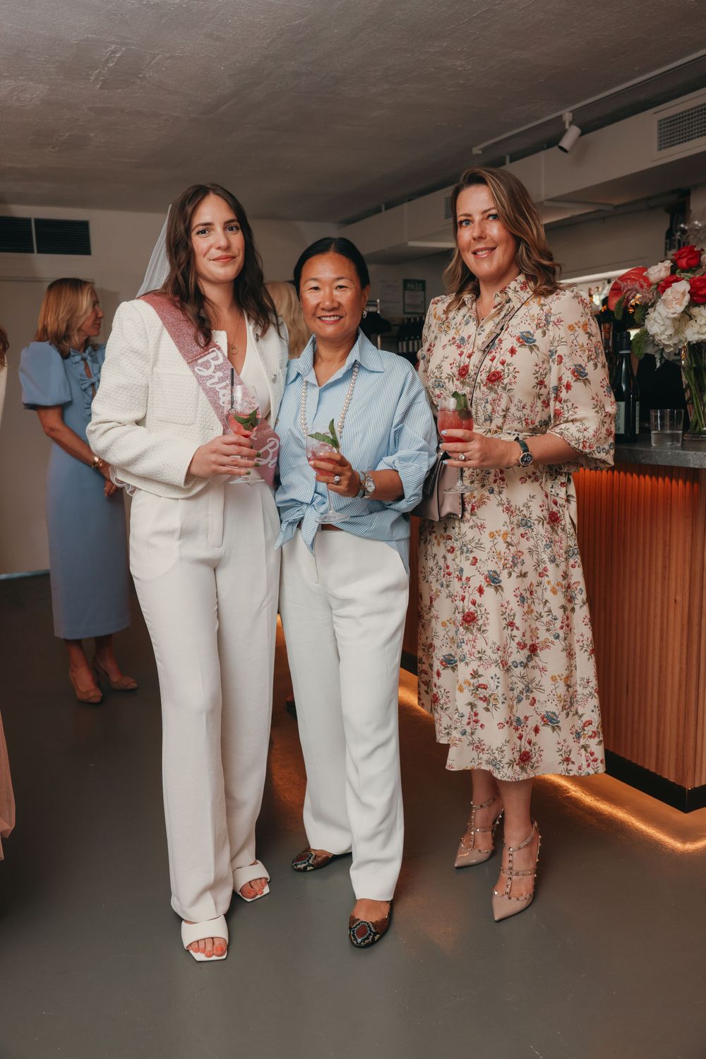 Three guests in elegant attire posing with drinks at social gathering.