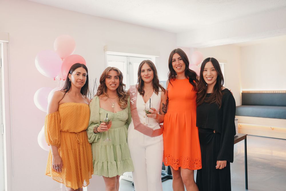 Five women in colorful dresses pose together at a celebration with pink decor.