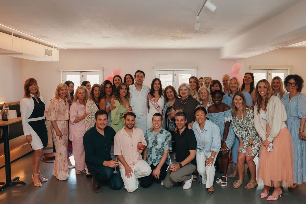 A large group of people wearing pastel and neutral colored outfits pose together for a group photo in a bright indoor space.