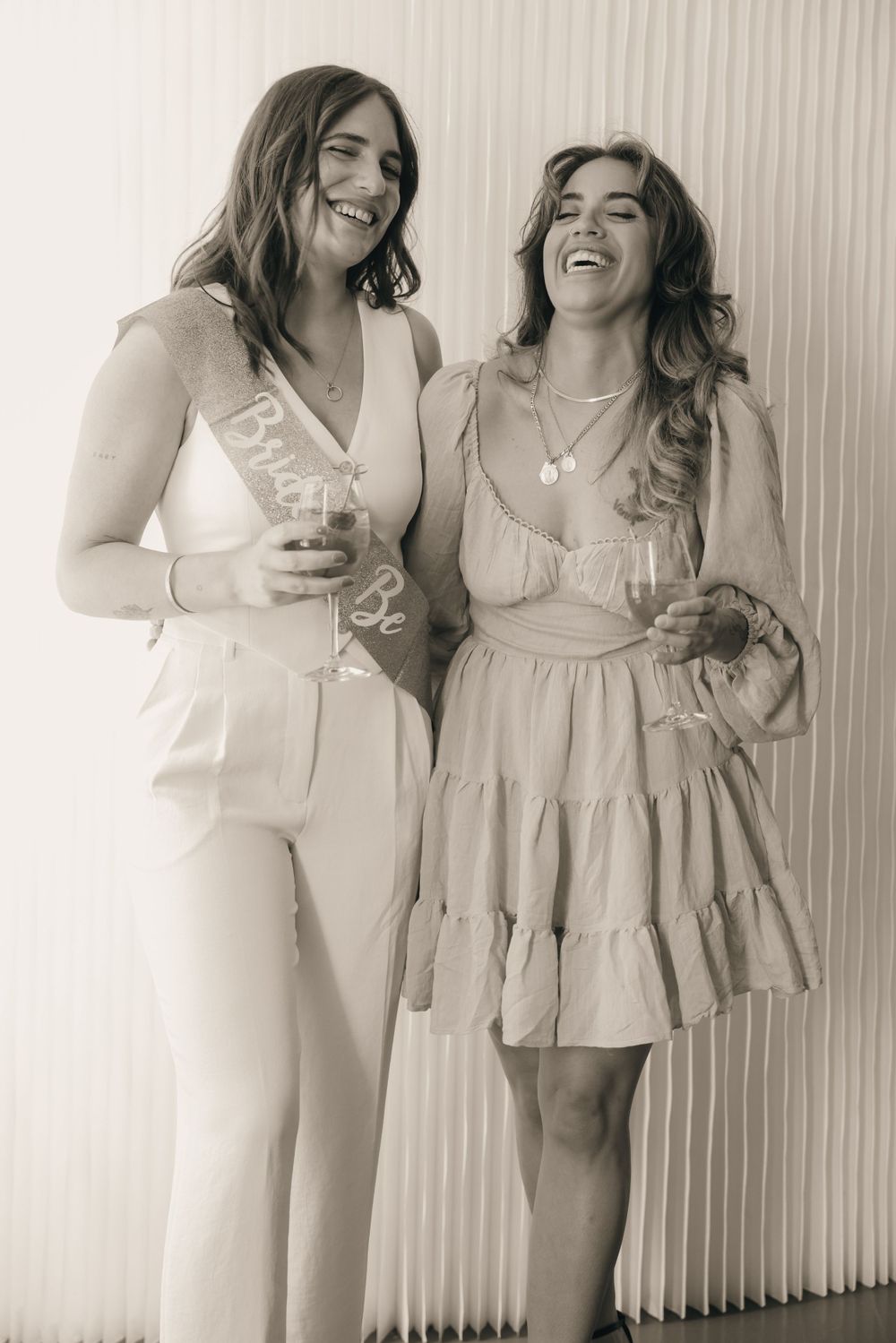Two people in light colored outfits share a laugh against a white textured wall.