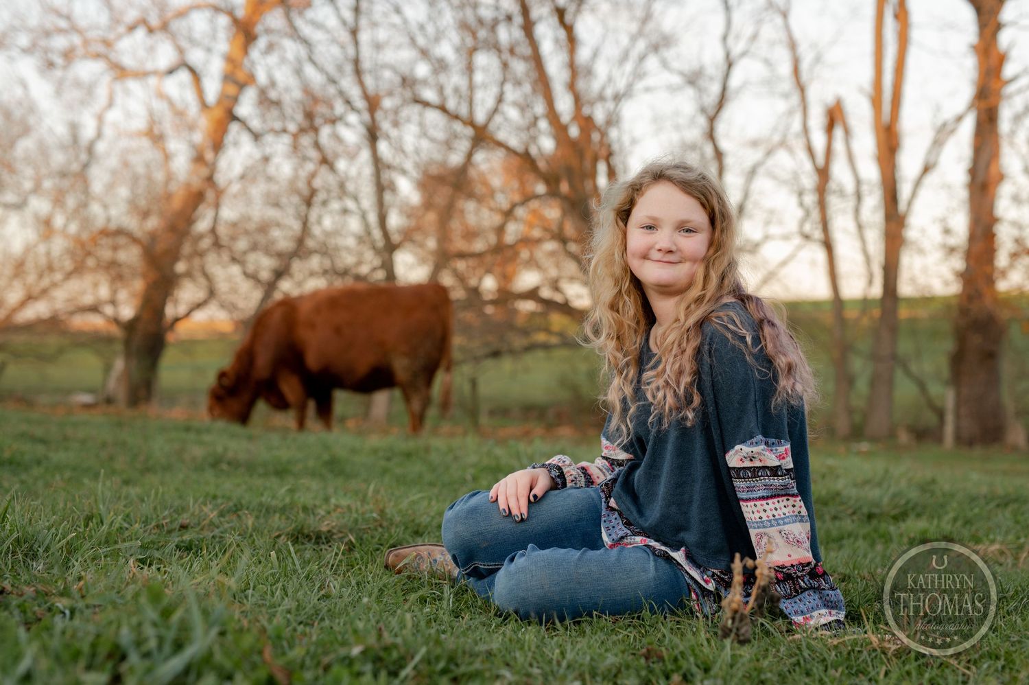 A person sits in a grassy field at sunset with a Highland cow grazing in the background.