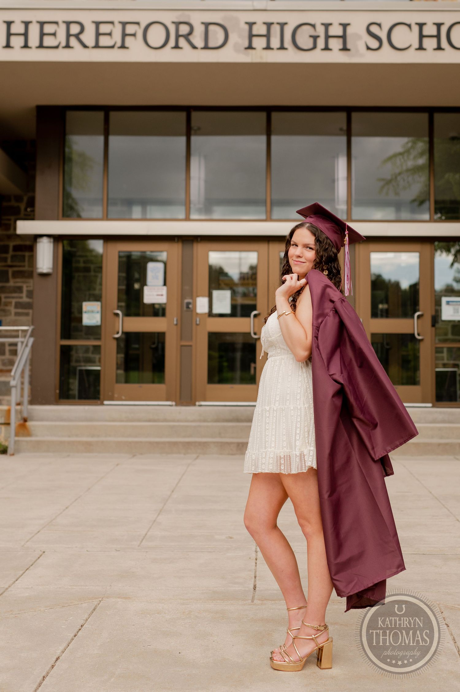 Graduate in white dress holds maroon graduation gown outside high school entrance.