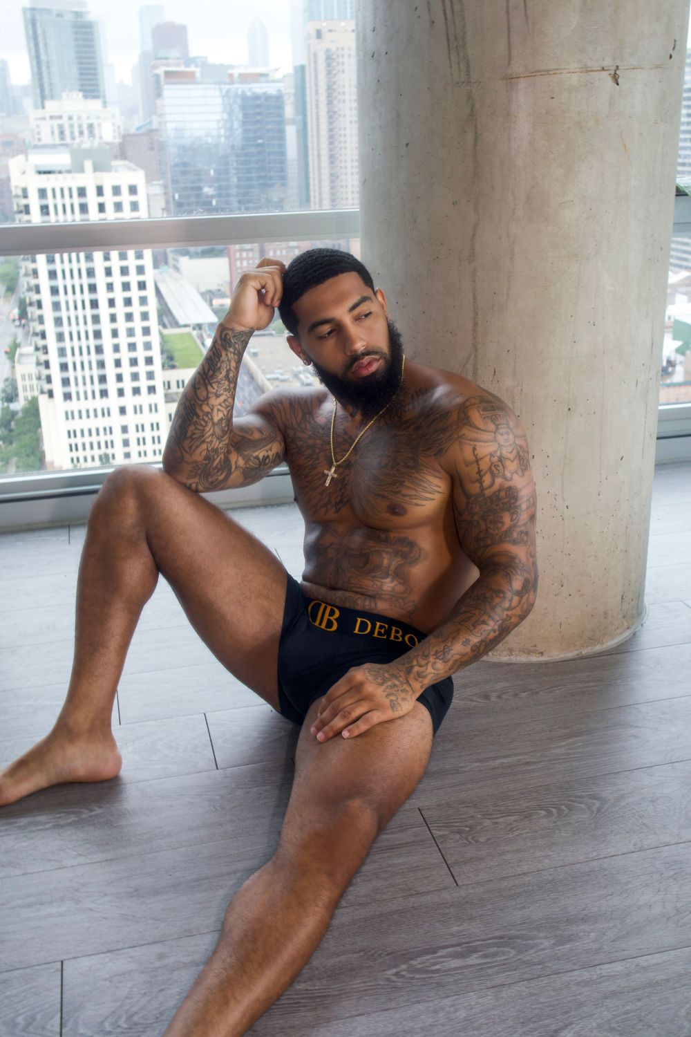 Athletic model wearing dark underwear poses near floor-to-ceiling windows with city skyline view in background.