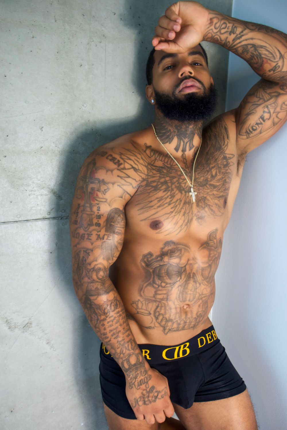Shirtless person with extensive tattoos poses against wall wearing black underwear with gold waistband.