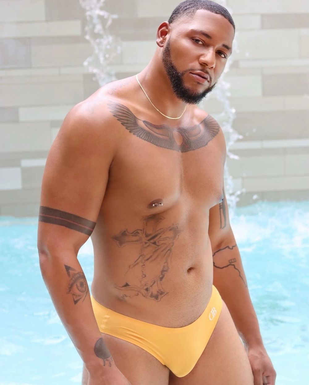 Athletic person in yellow swimwear posing by pool with tattoos and visible smoke in the background.