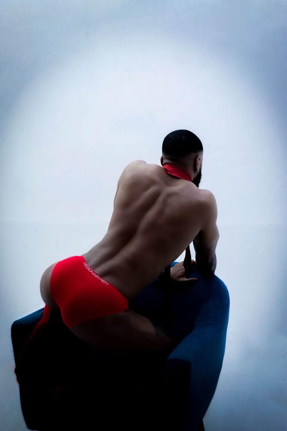 Artistic portrait showing a shirtless figure in red shorts sitting on a blue surface against a misty background.