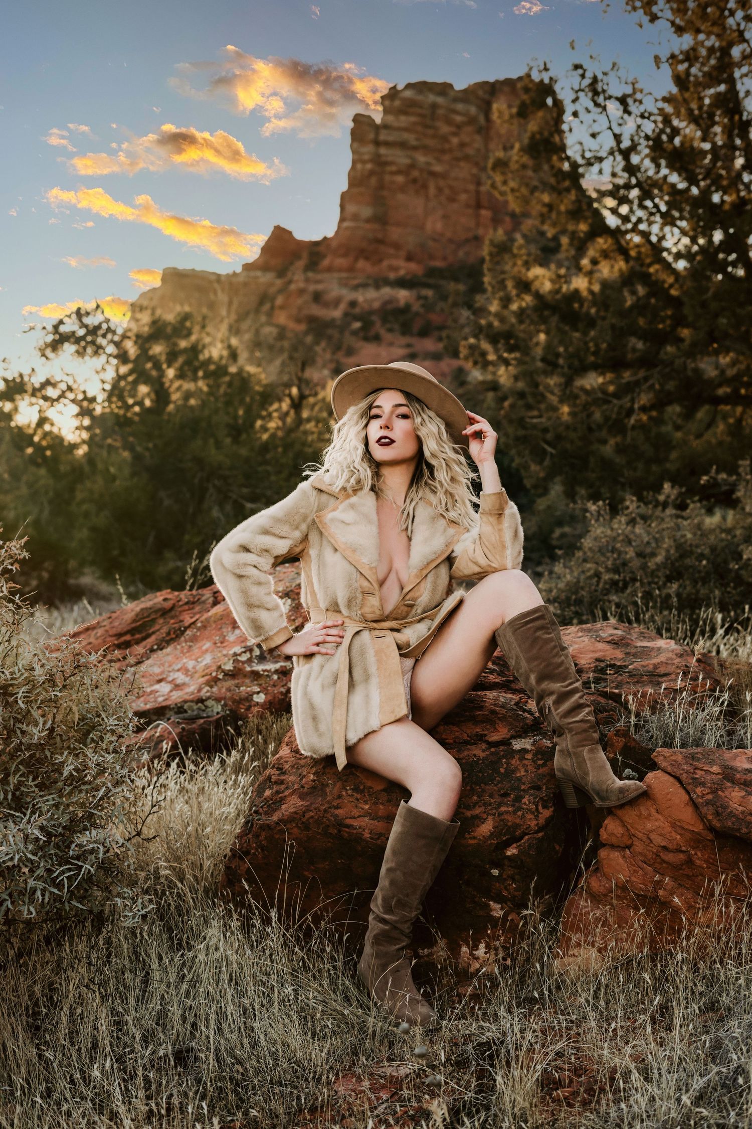 Fashion portrait in beige trench coat and boots against desert rock formation at sunset.