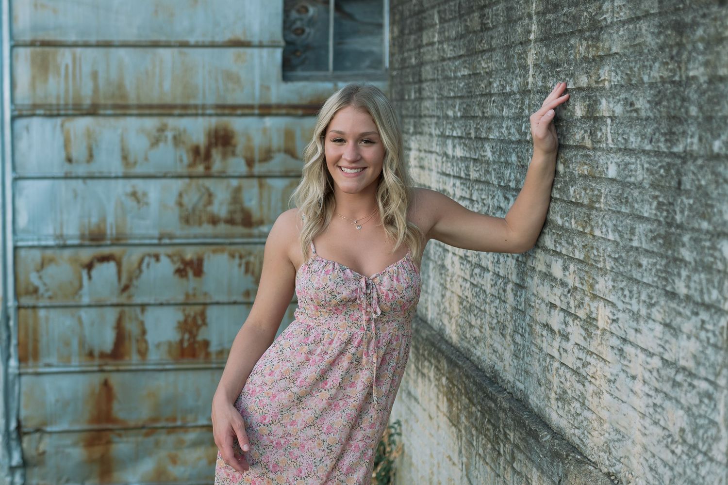 Woman in floral dress poses casually against rustic brick wall.