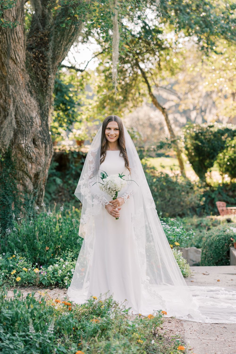 Bride holds white bouquet while standing in lush garden wearing long-sleeved lace wedding dress.