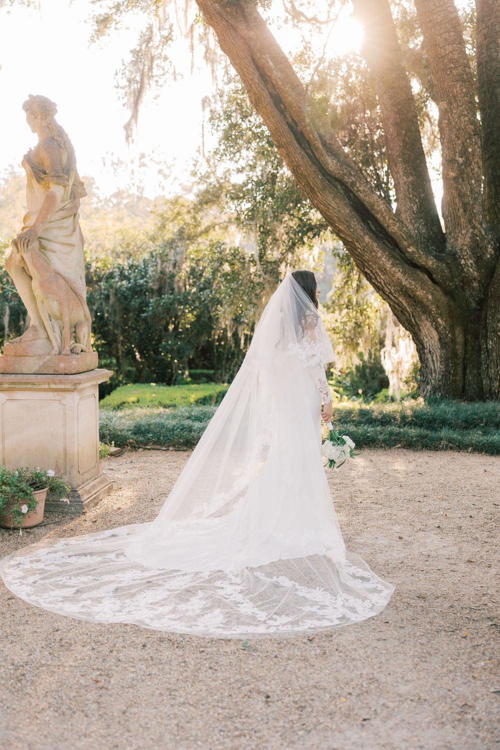 Cathedral length lace veil trails on garden path near classical statue.