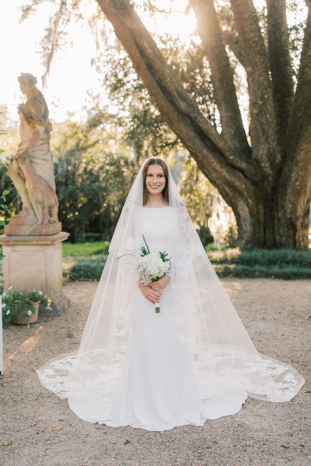 Elegant bridal portrait in a garden setting with a flowing cathedral veil and white bouquet beside a classical statue.