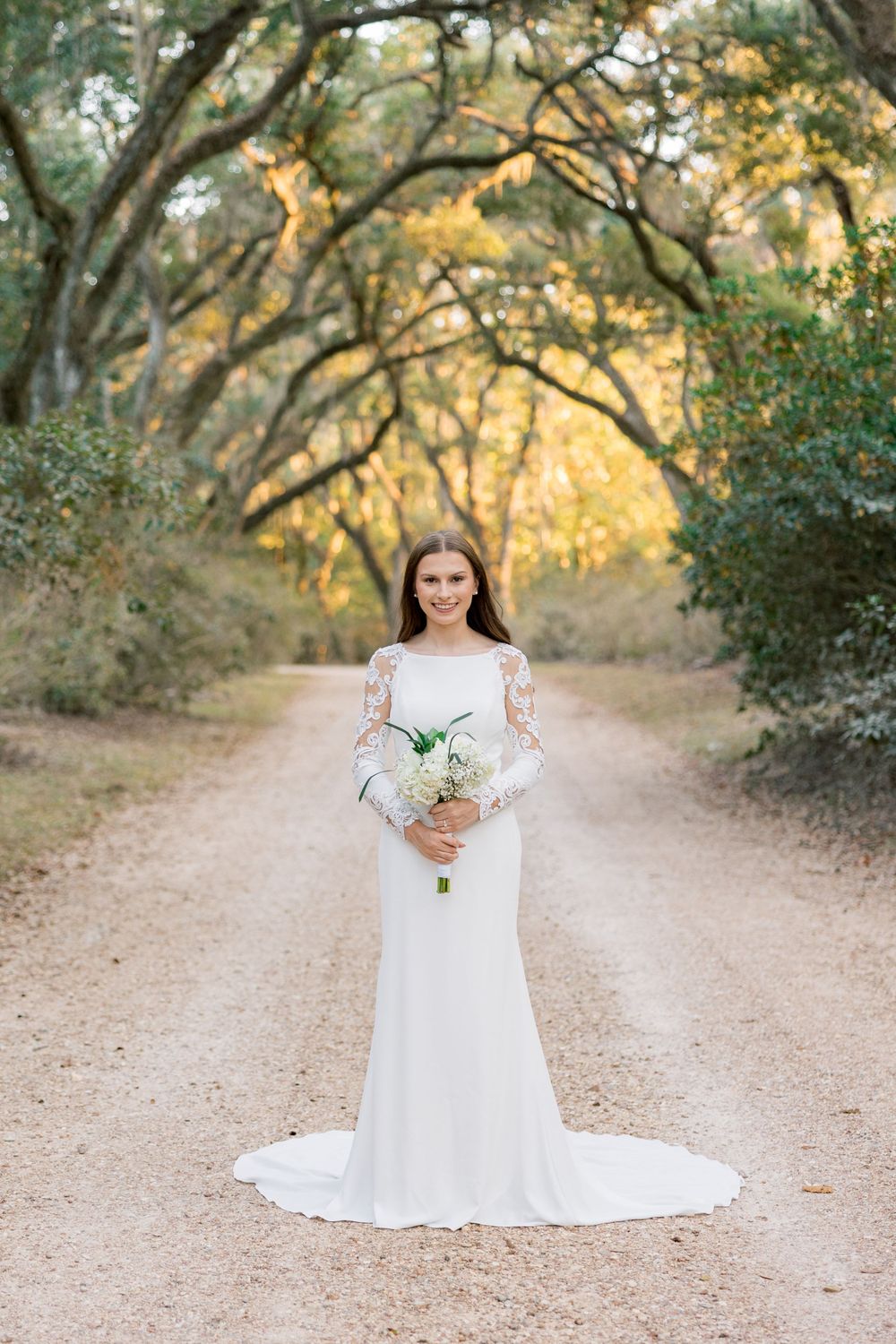 Elegant bridal portrait on a tree-lined path with white bouquet and fitted lace gown.