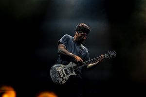 Guitarist performs on stage with a grey electric guitar against dark background with moody lighting.