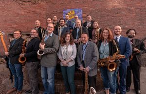 A group of jazz musicians with various instruments pose together against a brick wall background.