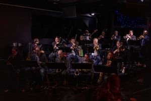A jazz orchestra performs on stage with dramatic lighting and multiple brass instruments visible.