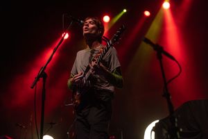 Musician playing electric guitar on dimly lit stage with red and yellow lighting effects.
