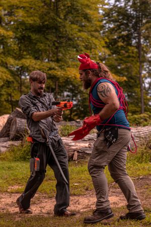 Two people in costume engage in LARP combat training with foam weapons in a forest setting.