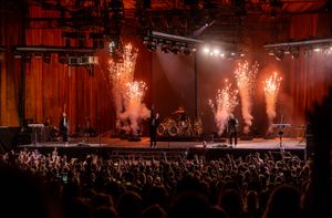 Rock concert stage with pyrotechnic effects and large crowd silhouetted in foreground.