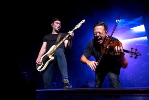 Musicians perform with guitars on stage with blue lighting effects.