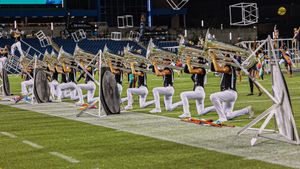 Marching band performers kneel in formation on a football field with silver flags at night.