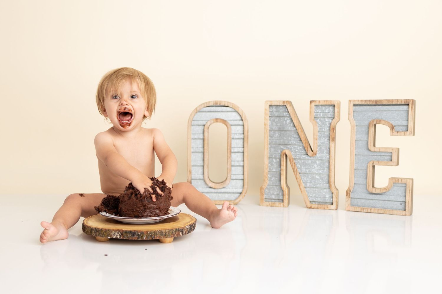 A first birthday cake smash photo setup with illuminated 'ONE' letters and chocolate cake on cream background.