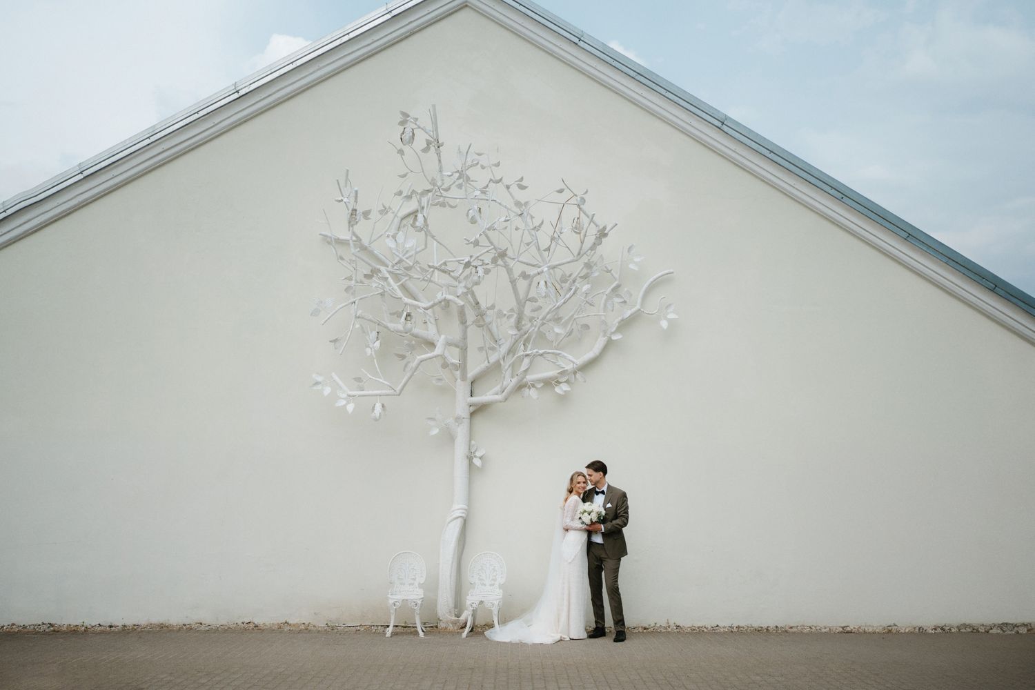 Wedding couple embraces against white wall with artistic tree sculpture.