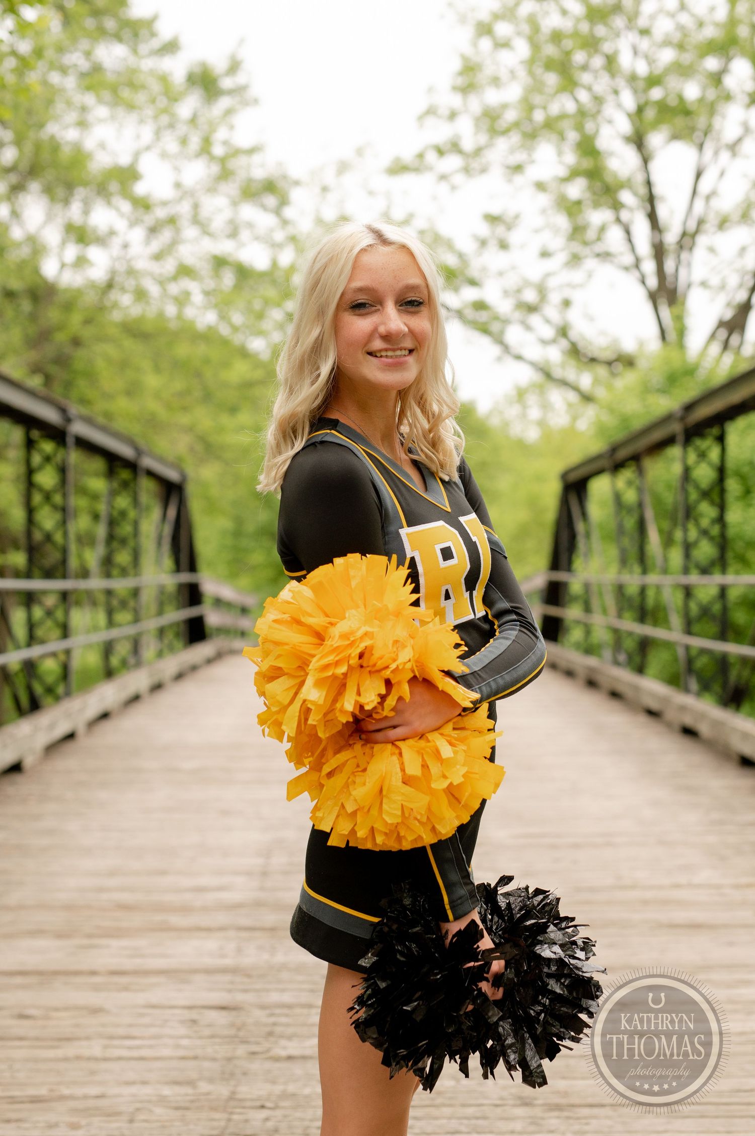 Cheerleader holds black and gold pom poms while posing on rustic wooden bridge in natural setting.
