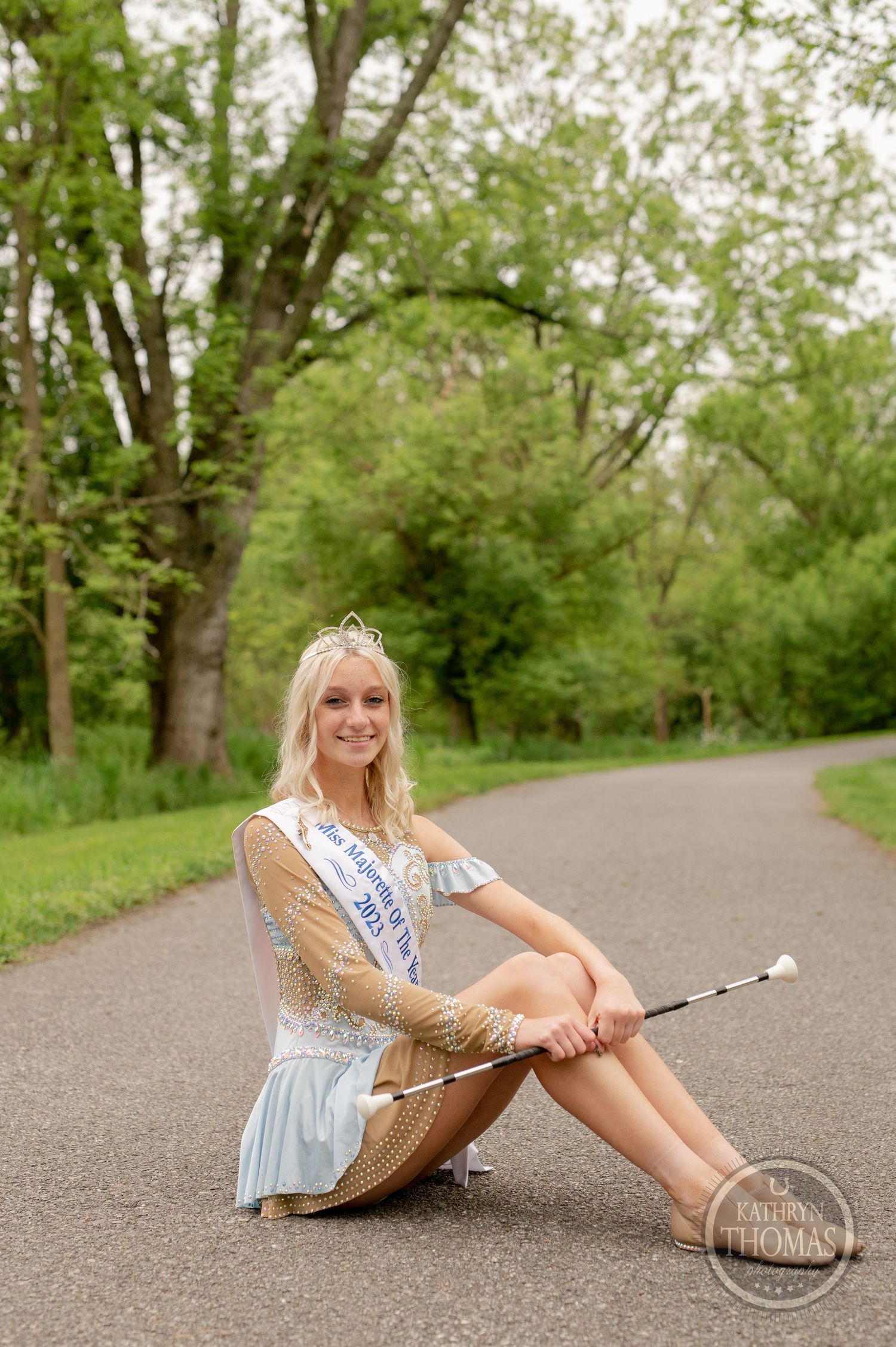Pageant contestant in sparkly dress sits on pathway holding baton with trees in background.