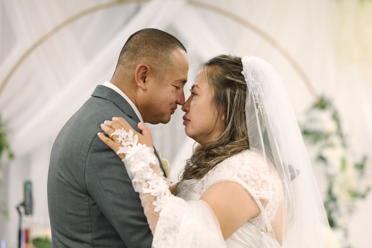 A bride and groom share a romantic kiss during their wedding ceremony under an arched backdrop with floral decorations.