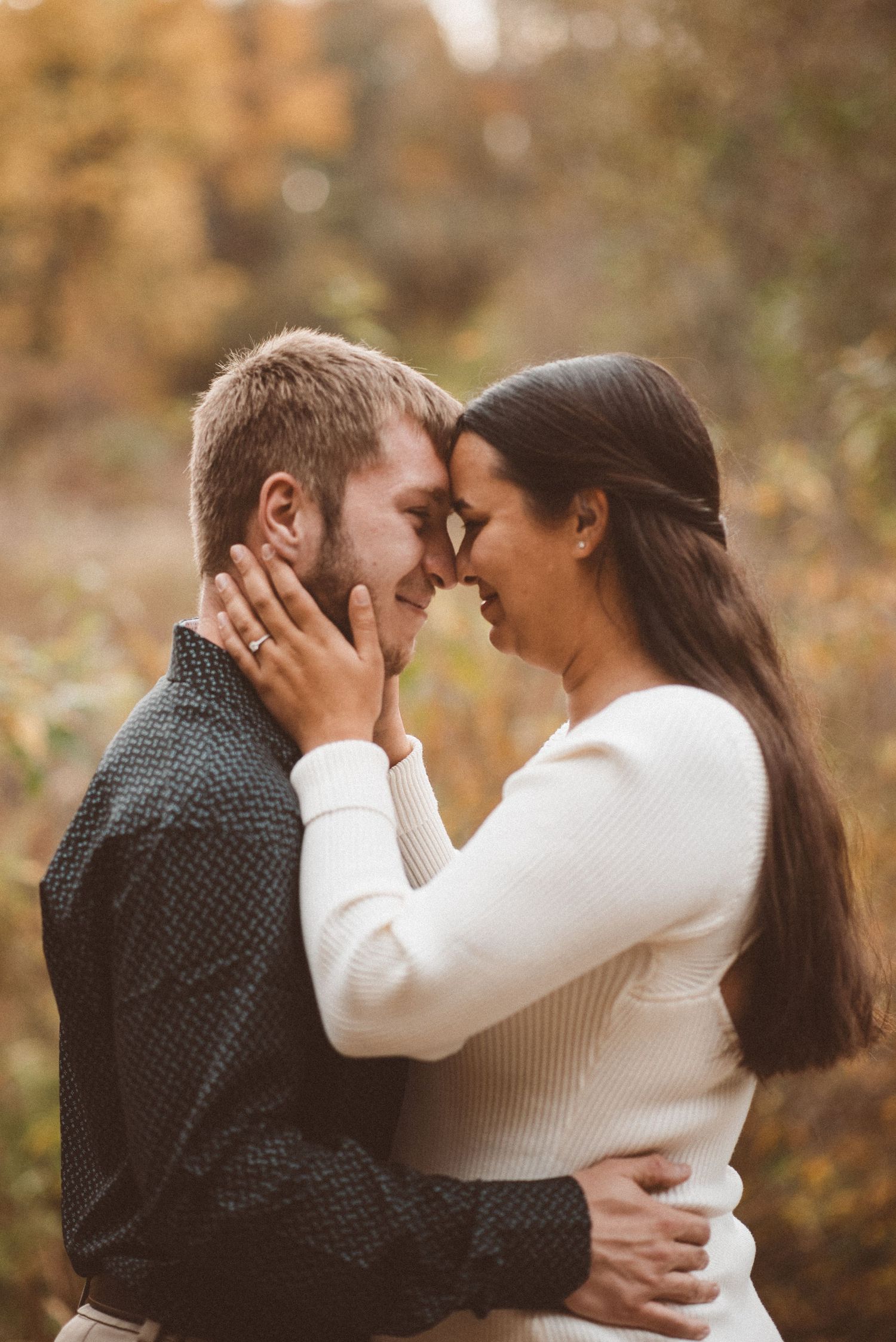 A couple facing each other for engagement pictures. The man's hands around her waist and her hands on his neck, showing off a shiny ring.