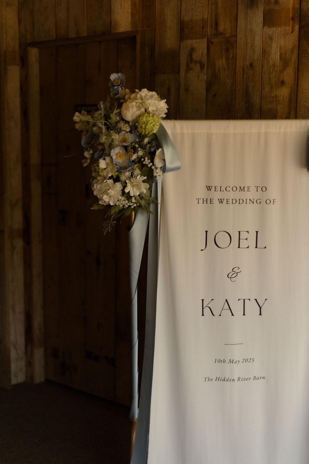 A wedding welcome sign for Joel and Katy with white hydrangeas and blue ribbon floral arrangement against wooden walls.