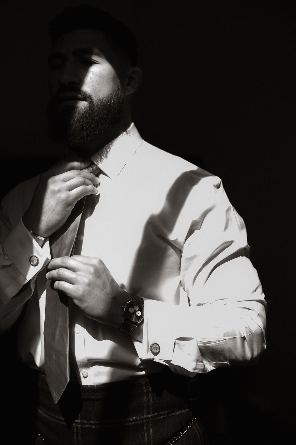 Black and white portrait of a bearded figure adjusting a tie while wearing a white dress shirt and formal attire.