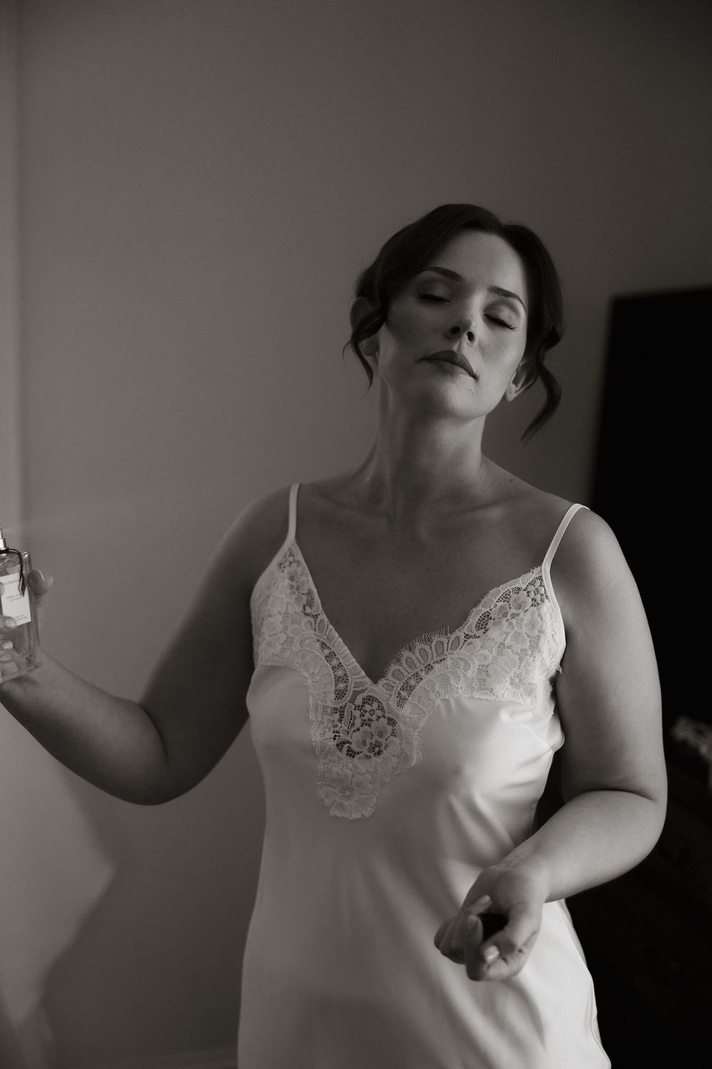 A black and white photograph shows a figure in a white lace-trimmed slip holding a perfume bottle in an intimate moment.