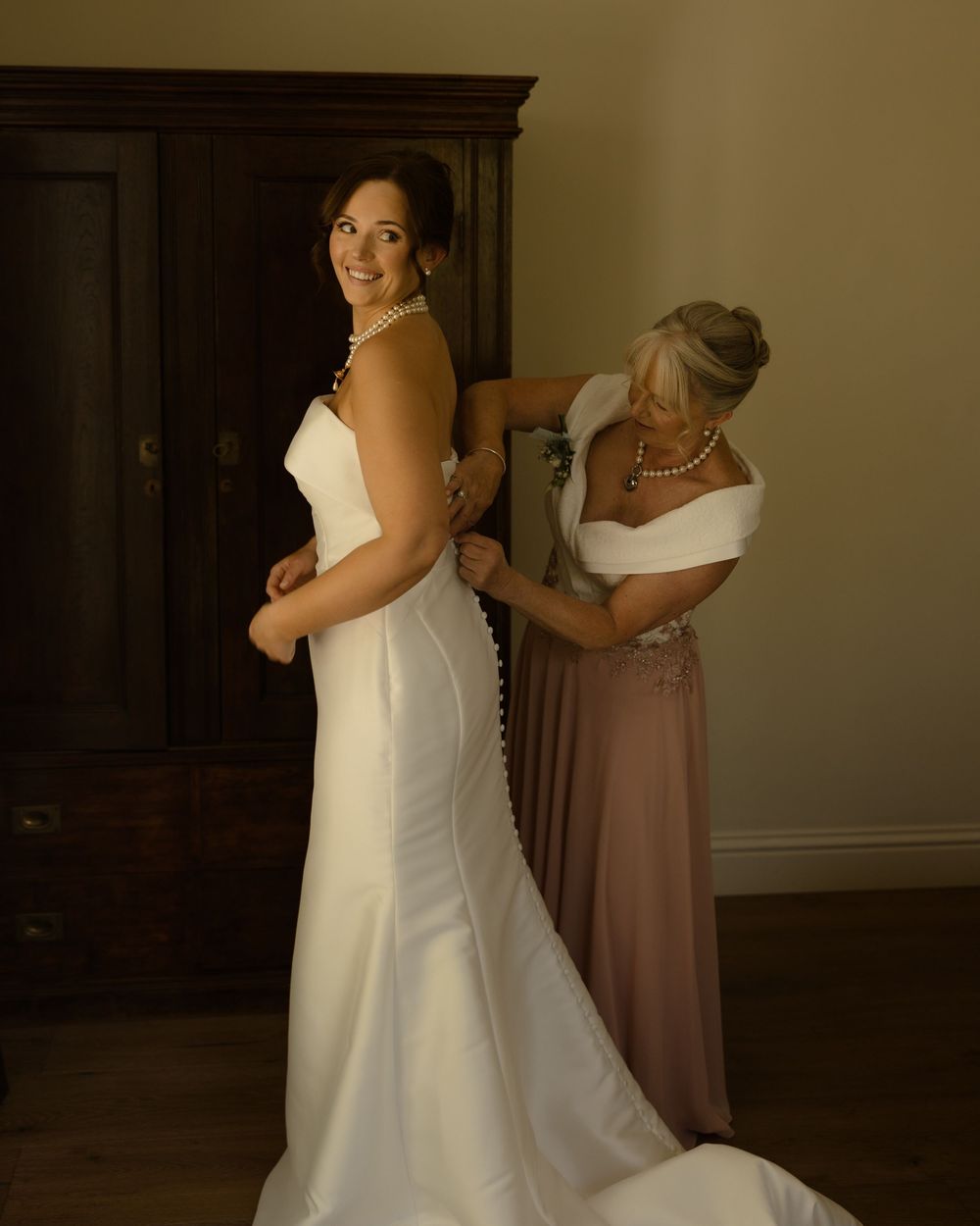 A person in a fitted white wedding gown gets help with the dress while standing in a warm-lit room with dark furniture.