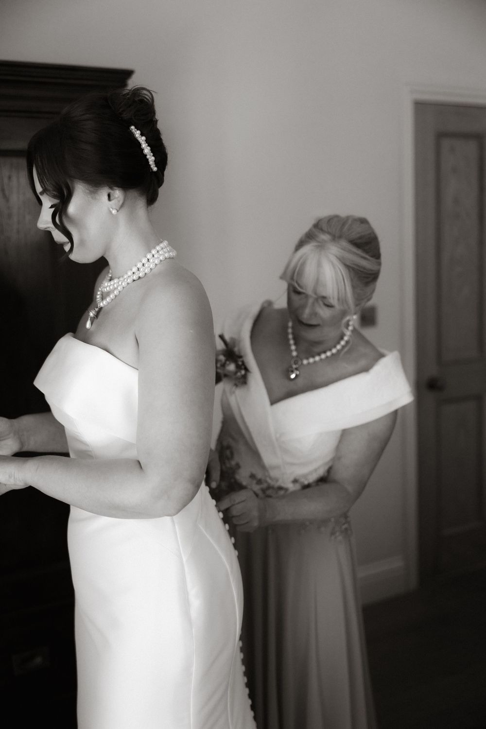 A black and white photo of wedding preparations showing someone getting help adjusting a white bridal gown.