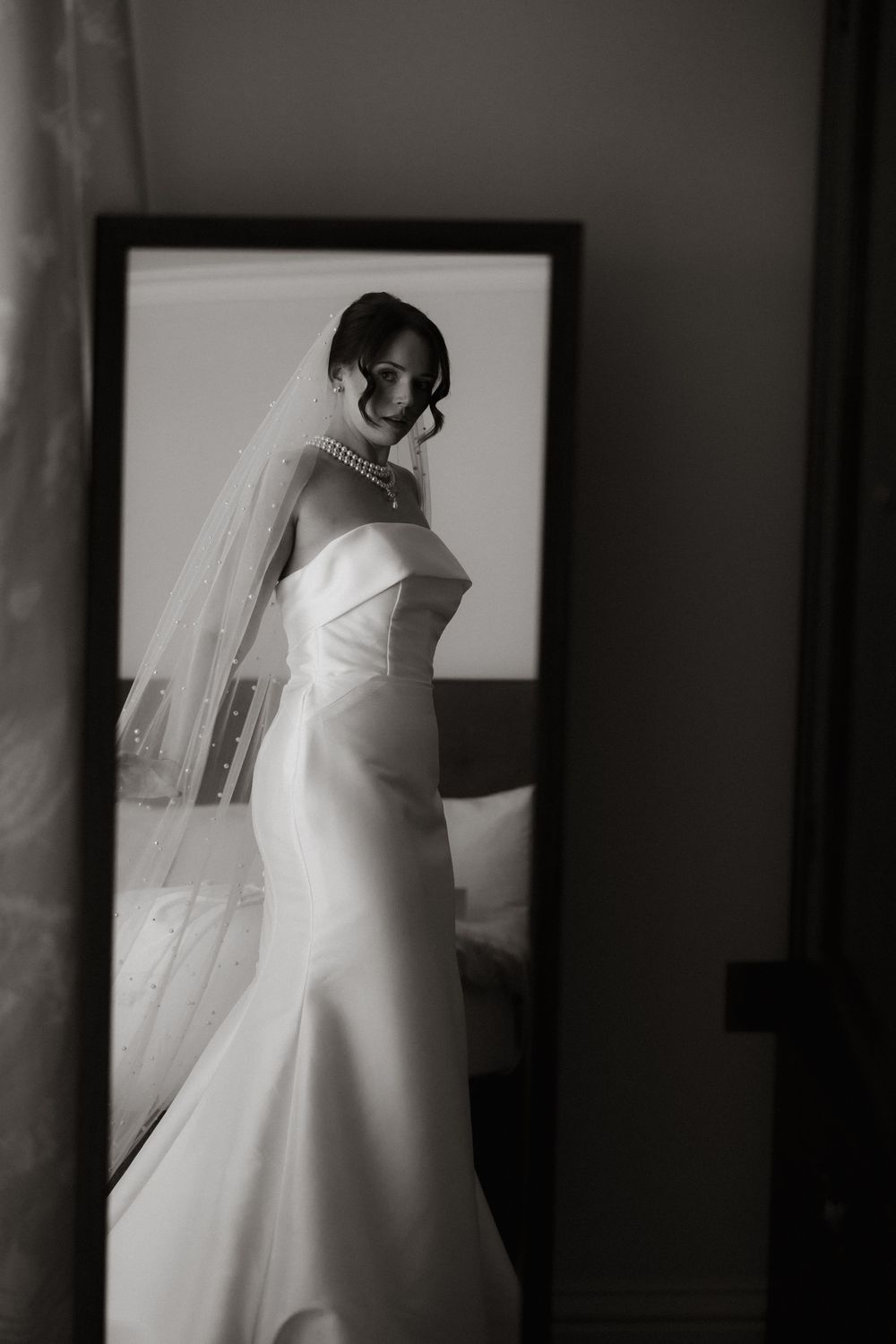 A bride in an elegant white wedding dress and long veil poses in a side profile in a monochrome photograph.