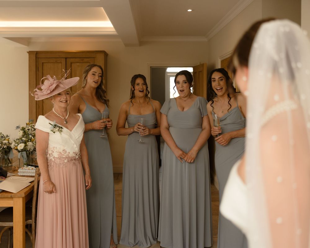 A bridal party in gray bridesmaid dresses shares a joyful moment before the wedding in an elegant indoor setting.