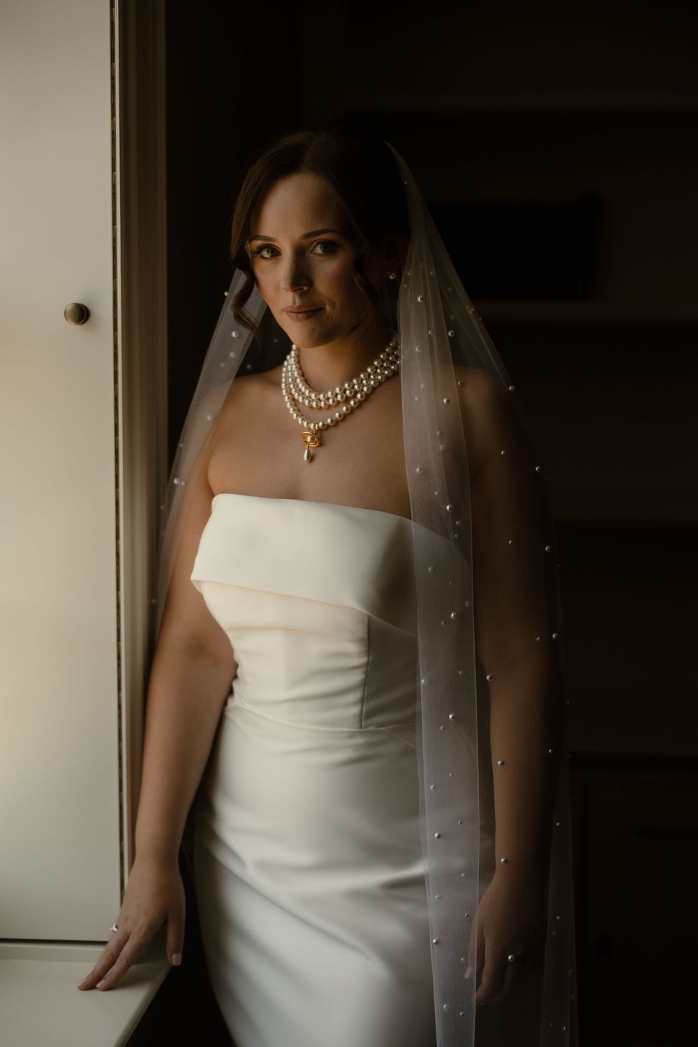 Elegant bridal portrait in white strapless gown with pearl necklace and veil standing by window in soft lighting.