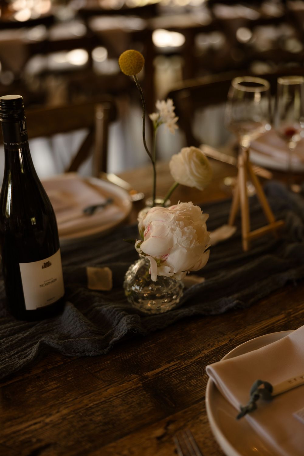 A rustic table setting with wine bottle and delicate white rose in a small vase against a dark wooden surface.