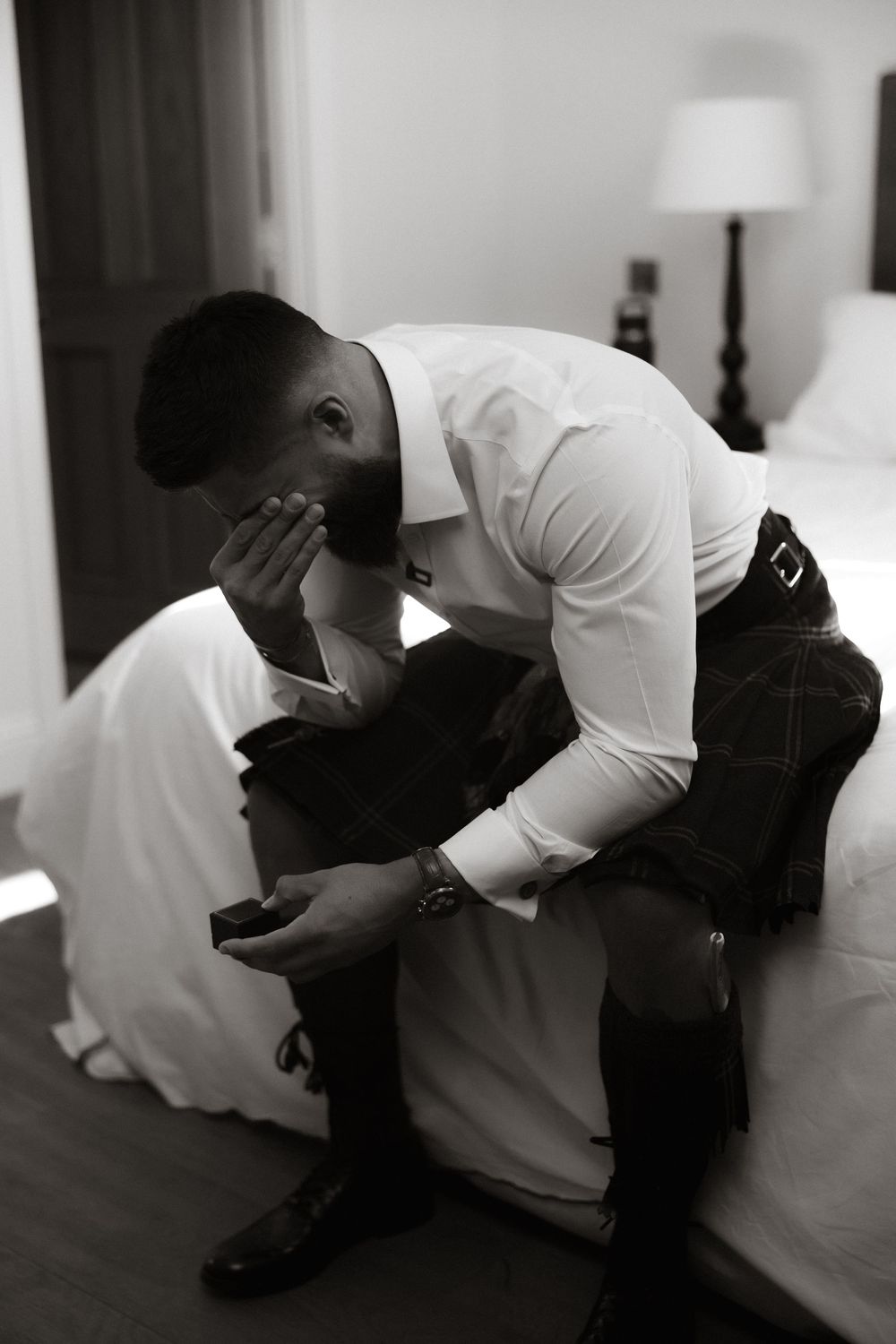 Black and white photo of person in formal attire sitting on bed with head in hands showing contemplative moment.