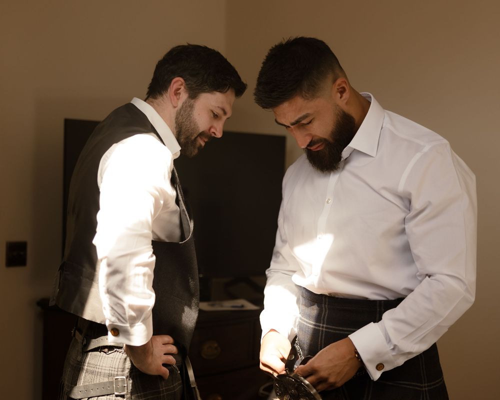 Two people in white dress shirts adjusting their outfits while getting ready in a dimly lit room.
