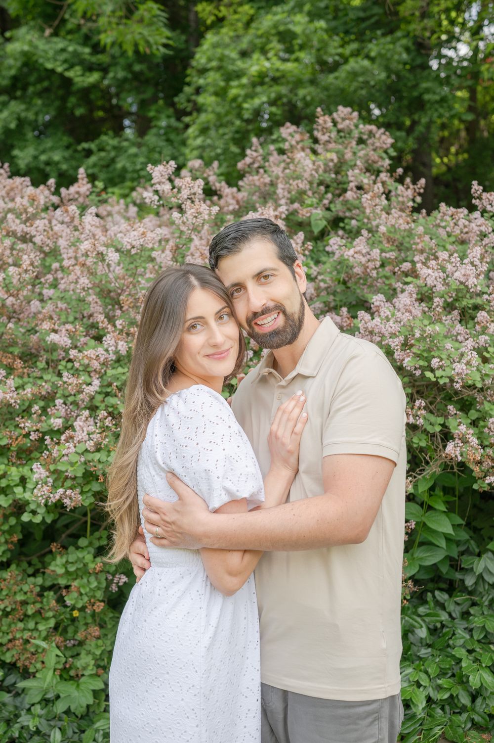 A romantic couple embraces in front of blooming pink flowers and green foliage on a sunny spring day.