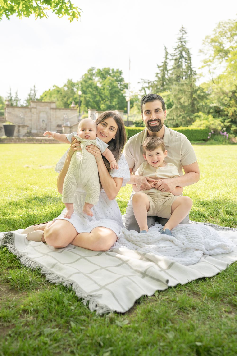 A family sits together on a blanket in a sunny garden with green grass and trees in the background.